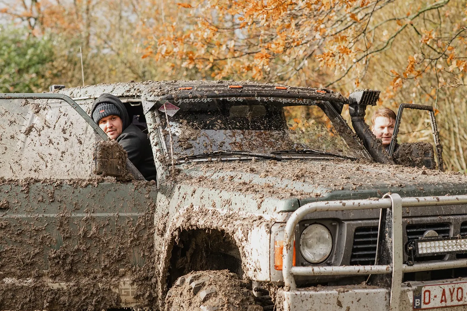 Twee deelnemers kijken lachend uit een terreinwagen die onder de modder zit